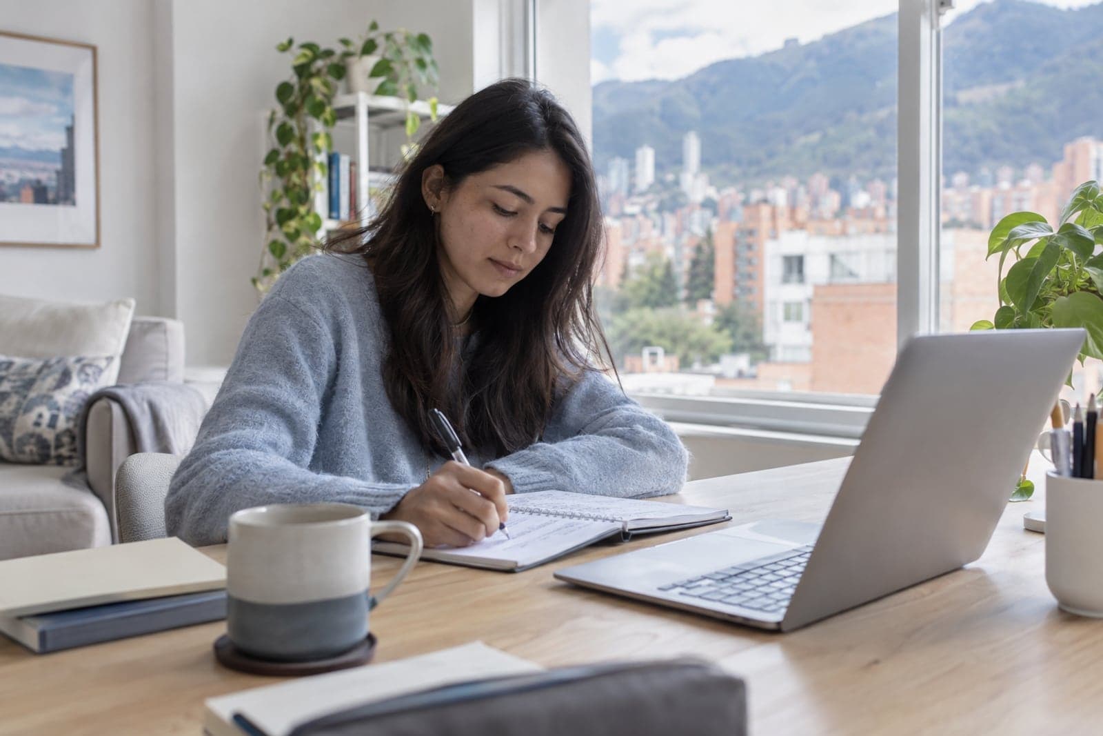 A student takes notes during an online course at her desk by a window with a city view.